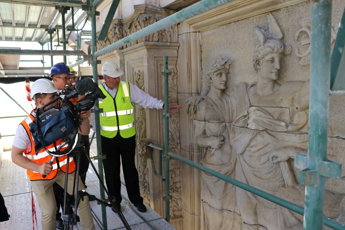 Peter Lochert, in a hi vis vest and helmet, points to details in Parliament House's facade. Two men from Channel 9 news record using a camera on a tripod.