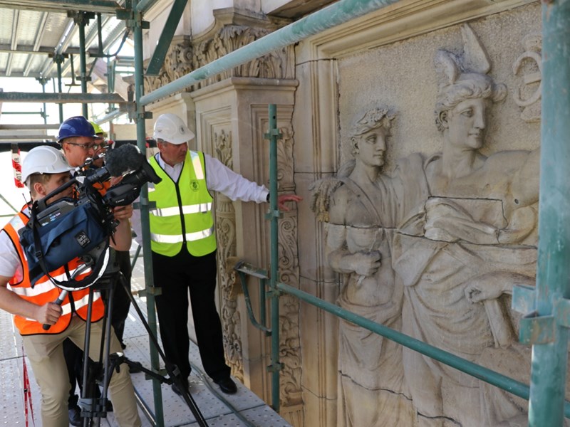 Peter Lochert, in a hi vis vest and helmet, points to details in Parliament House's facade. Two men from Channel 9 news record using a camera on a tripod.